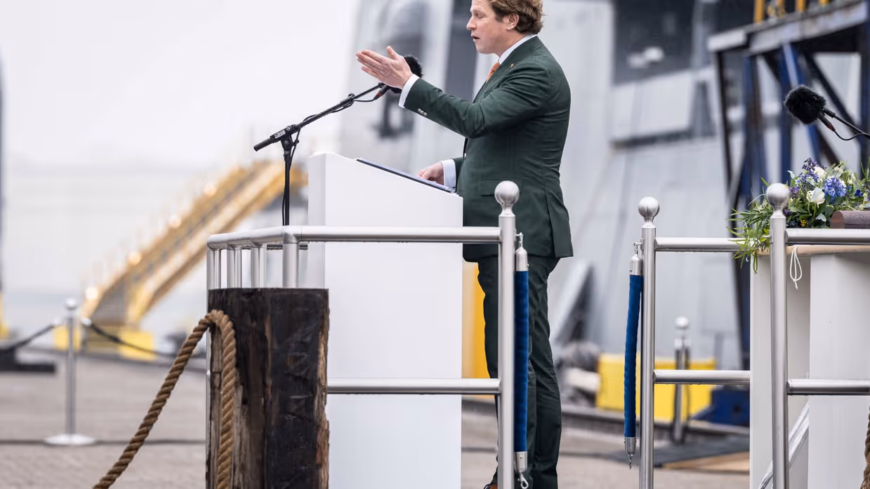 Staatssecretaris Gijs Tuinman in groen pak achter katheder op kade. Marineschip is vaag op de achtergrond zichtbaar.