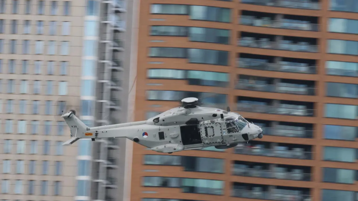 Een lichtgrijze NH90-maritieme gevechtshelikopter vliegt op snelheid langs hoge flatgebouwen. 