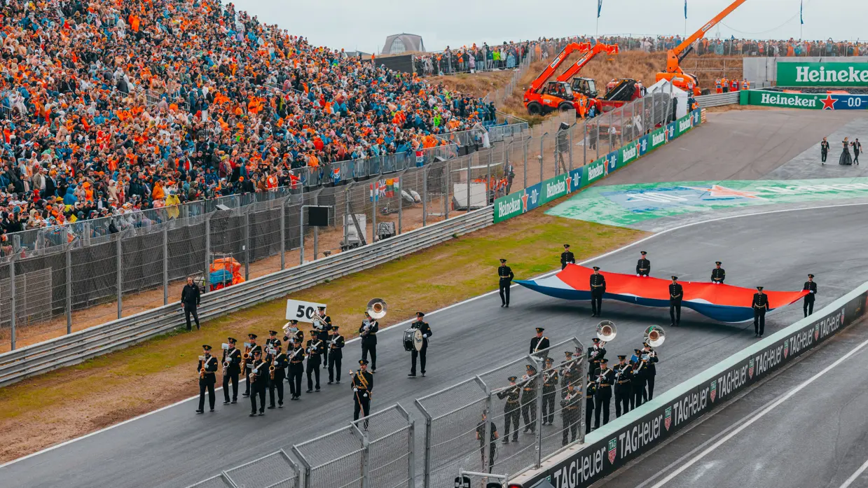 Het Orkest Koninklijke Luchtmacht speelt muziek midden op de racebaan. Achter het orkest houden acht militairen een grote vlag vast die bijna even breed is als het wegdek. Naast de racebaan staat een tribune vol publiek.