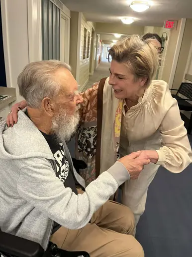 Een vrouw en oudere man zitten glimlachend naast elkaar. Zij houdt zijn hand vast. Achter hen staat een bordje met "Blessed". Warm, gezellig interieur.