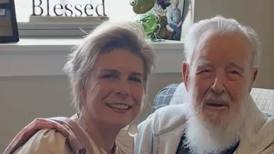 Een vrouw en oudere man zitten glimlachend naast elkaar. Zij houdt zijn hand vast. Achter hen staat een bordje met "Blessed". Warm, gezellig interieur.