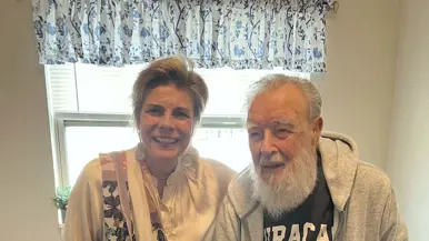 Een vrouw en oudere man zitten glimlachend naast elkaar. Zij houdt zijn hand vast. Achter hen staat een bordje met "Blessed". Warm, gezellig interieur.