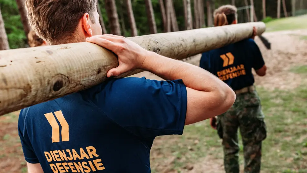 2 jongeren met shirt Dienjaar Defensie sjouwen een boomstam.
