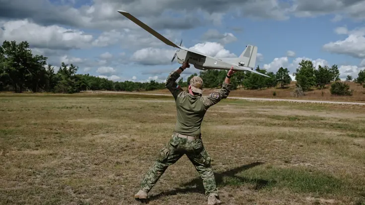 Militair in open veld met grote drone in zijn armen boven z'n hoofd.