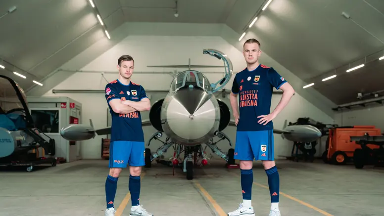 Twee mannen staan in een voetbaltenue naast een oude straaljager in een zaal.