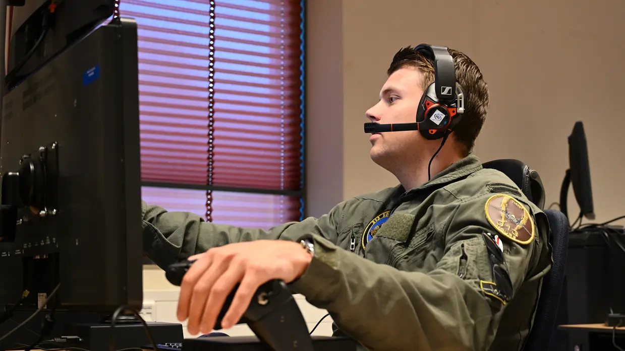 Vlieger met headset op achter een bureau met computerscherm, zijn linkerhand rust op een zwarte hendel.