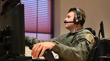 Vlieger met headset op achter een bureau met computerscherm, zijn linkerhand rust op een zwarte hendel.