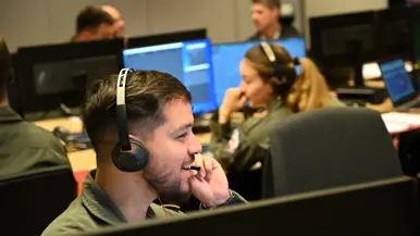 : Militair met headset op zit achter een beeldscherm en spreekt lachend door zijn microfoon. Op de achtergrond meerdere militairen aan bureaus met schermen.