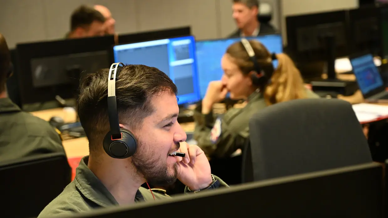 : Militair met headset op zit achter een beeldscherm en spreekt lachend door zijn microfoon. Op de achtergrond meerdere militairen aan bureaus met schermen.