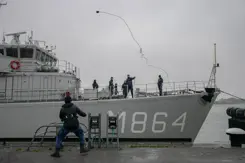 Marineschip legt aan bij kade. Militair gooit touw naar collega op kade.