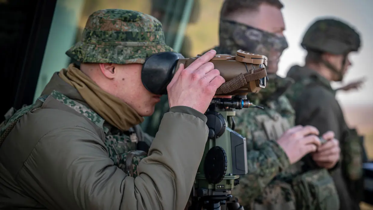 Drie militairen houden overzicht op de werkzaamheden. De man op voorgrond kijkt door een stel verrekijkers.