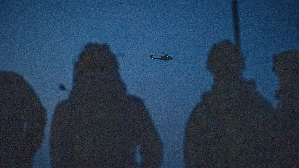 Vier militairen staan met hun rug naar de camera 's avonds klaar, in de lucht verderop is een helikopter aan het vliegen.