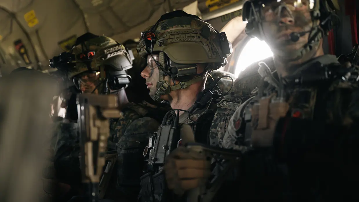 Gewapende parachutisten met helmen en communicatiemiddelen zitten geconcentreerd in een donker transportvliegtuig vlak voor een nachtelijke airborne-operatie.	 