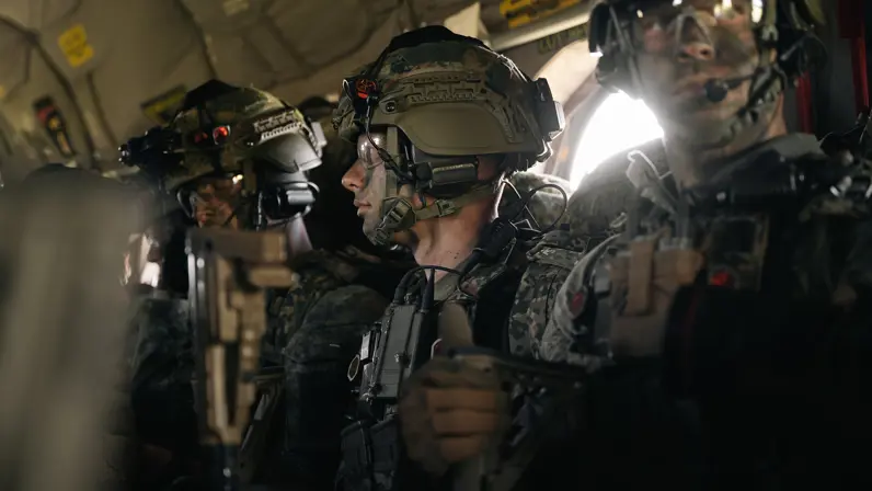 Gewapende parachutisten met helmen en communicatiemiddelen zitten geconcentreerd in een donker transportvliegtuig vlak voor een nachtelijke airborne-operatie.	 