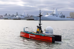 Een varend rood vaartuig dat op afstand bestuurbaar is met marineschepen op de achtergrond.