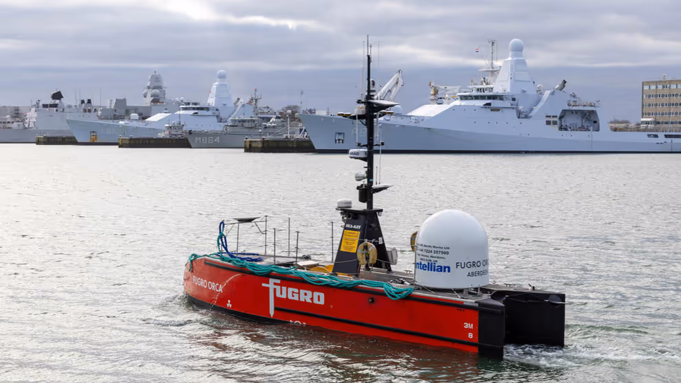 Een varend rood vaartuig dat op afstand bestuurbaar is met marineschepen op de achtergrond.