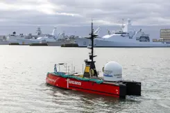 Een varend rood vaartuig dat op afstand bestuurbaar is met marineschepen op de achtergrond.