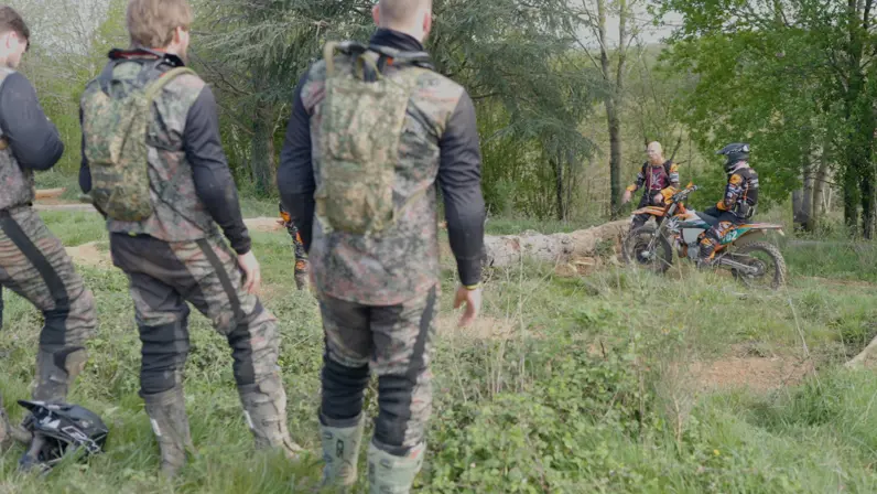 Een man op de motor wordt toegesproken door een andere man in motorpak. Op de voorgrond staan drie militairen met hun rug naar de camera.