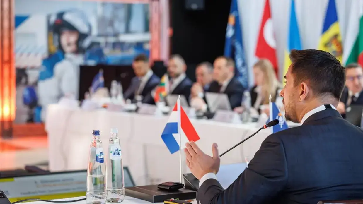 Een Nederlandse afgevaardigde spreek zijn collega’s toe vanachter een tafel met daarop een Nederlands vlaggetje.