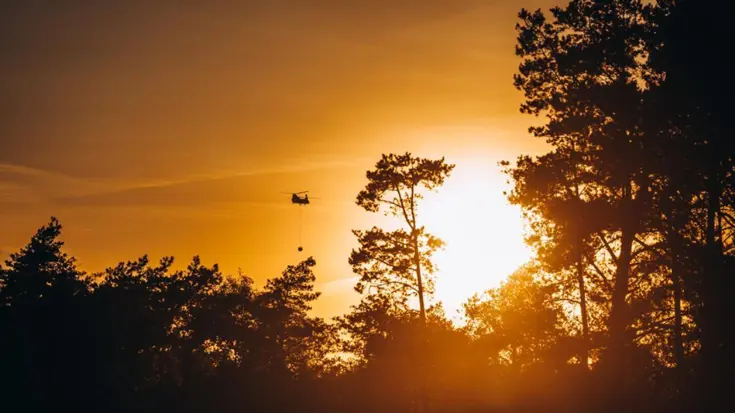 Een Chinook met een Bambi Bucket eronder vliegt boven een bosbrand. 
