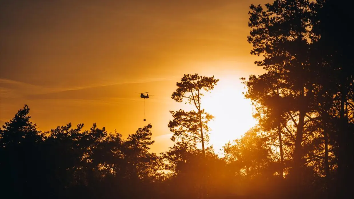 Een Chinook met een Bambi Bucket eronder vliegt boven een bosbrand. 