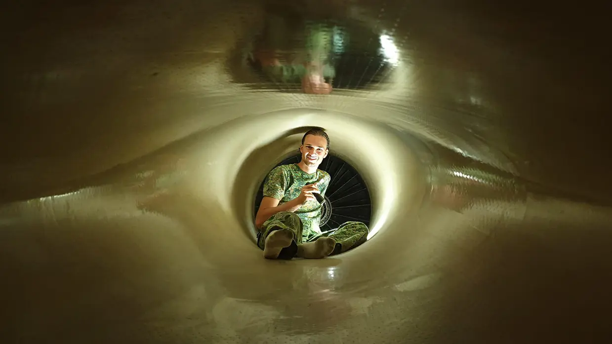Man in groen camouflagepak zit glimlachend in een smalle, verlichte tunnel met een fietswiel op de achtergrond.
