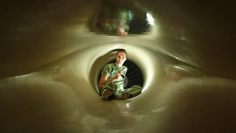Man in groen camouflagepak zit glimlachend in een smalle, verlichte tunnel met een fietswiel op de achtergrond.