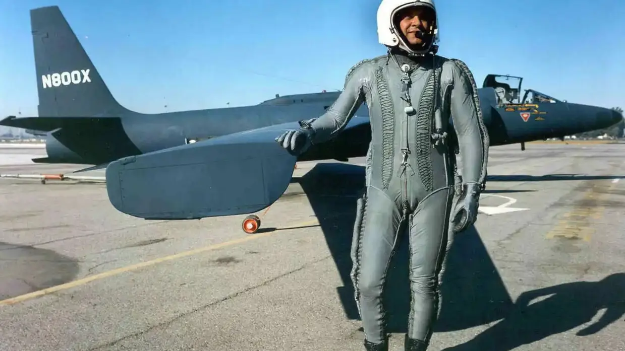 Een persoon in een strak, grijs pilotenpak met witte helm, staat glimlachend voor een donker, futuristisch ogend vliegtuig op een vliegveld. Het is mooi weer.
