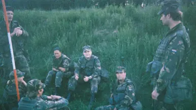 Een jonge Tim Nelissen in gevechtstenue zittend in het hoge gras samen met zes andere militairen, die op hem heen zitten en staan. Tim steekt beide duimen op richting de camera.