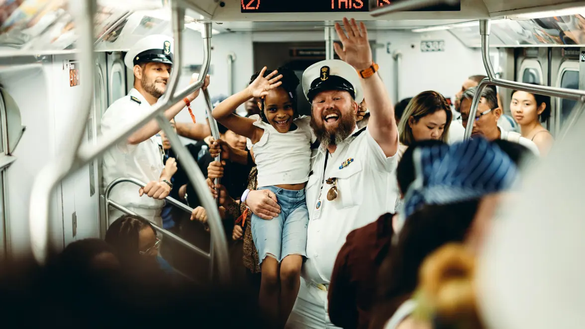  Een marineman in de metro, die een meisje op zijn arm heeft. Beiden zwaaien naar de camera. 