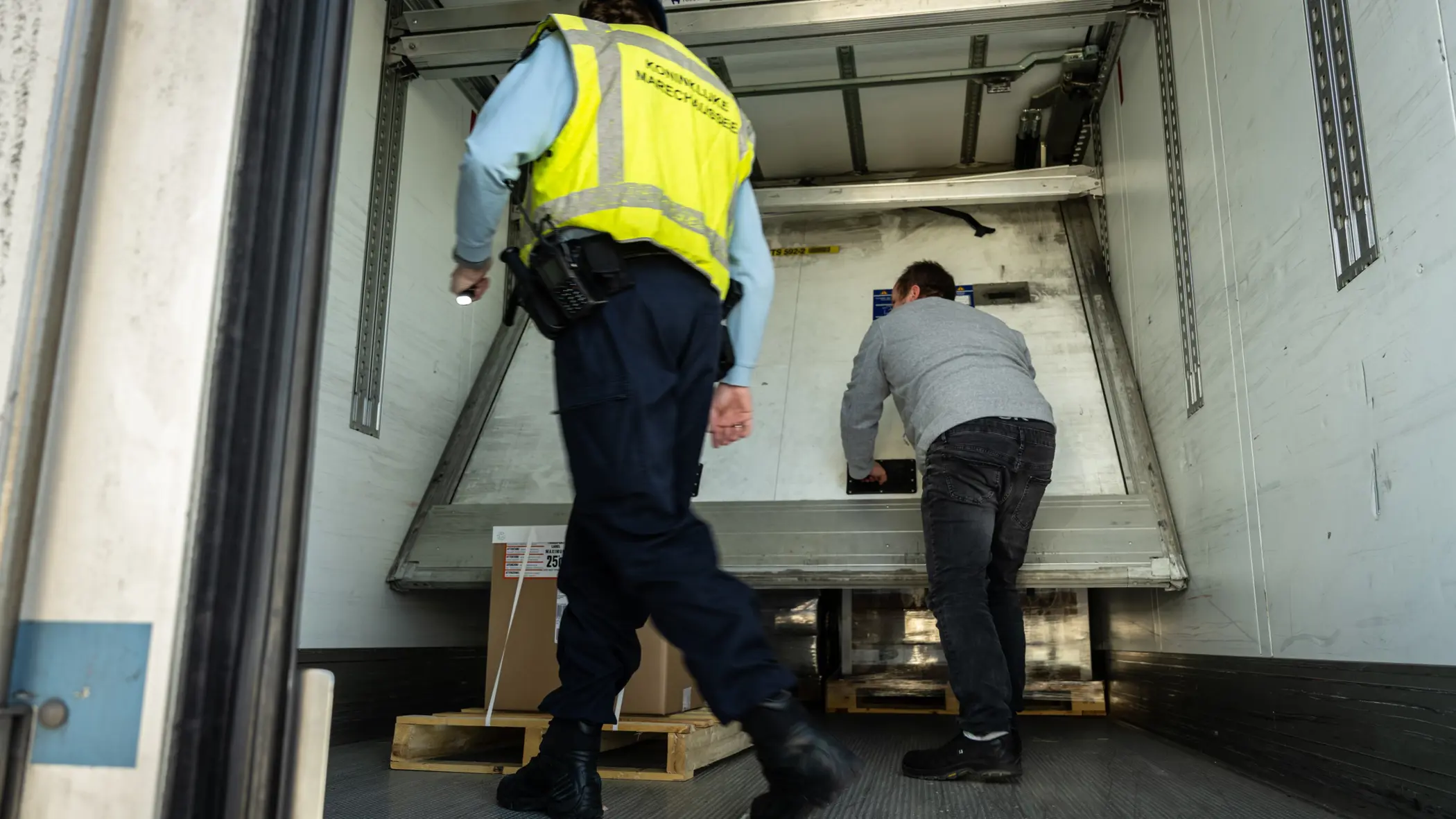 Een chauffeur schuift in een oplegger een paneel, weg dat voor de lading zit. Een marechaussee kijkt toe.