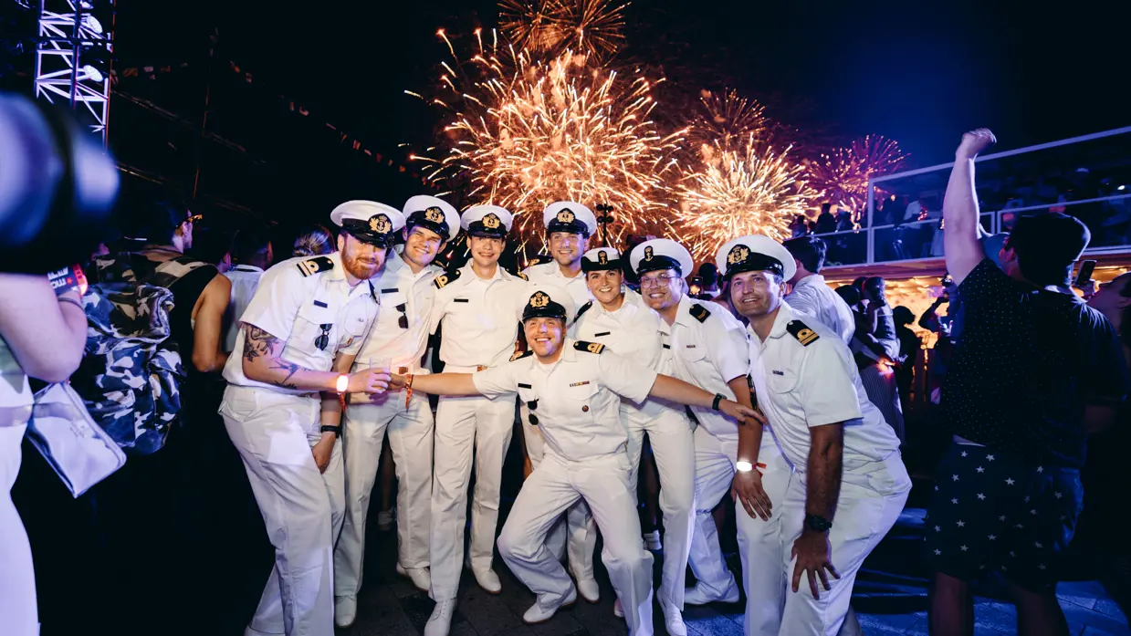 een poserend groepje opvarenden in wit uniform met op de achtergrond vuurwerk.  