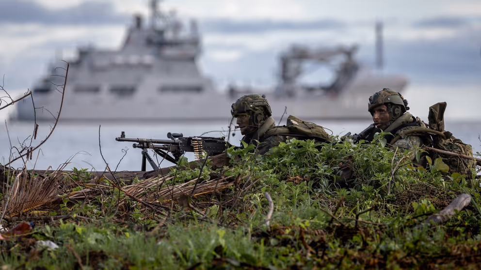 Mariniers liggen op de grond met op de achtergrond een varend marineschip.
