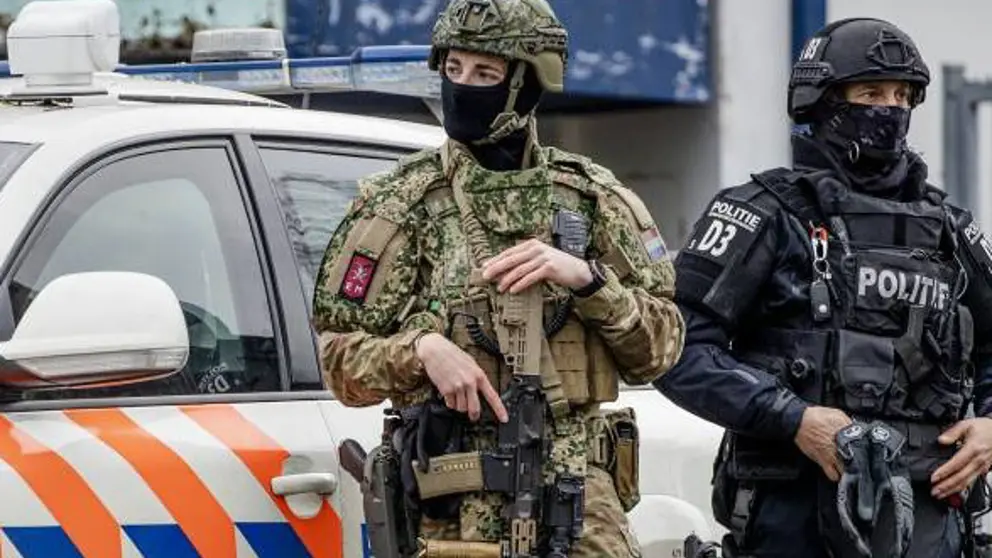 En gewapende, gemaskerde militair staat voor een politieauto en naast een politieman met gezichtsbedekking.