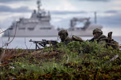 Mariniers liggen op de grond met op de achtergrond een varend marineschip.