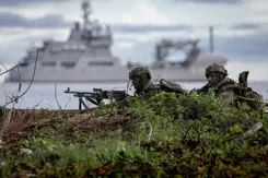 Mariniers liggen op de grond met op de achtergrond een varend marineschip.