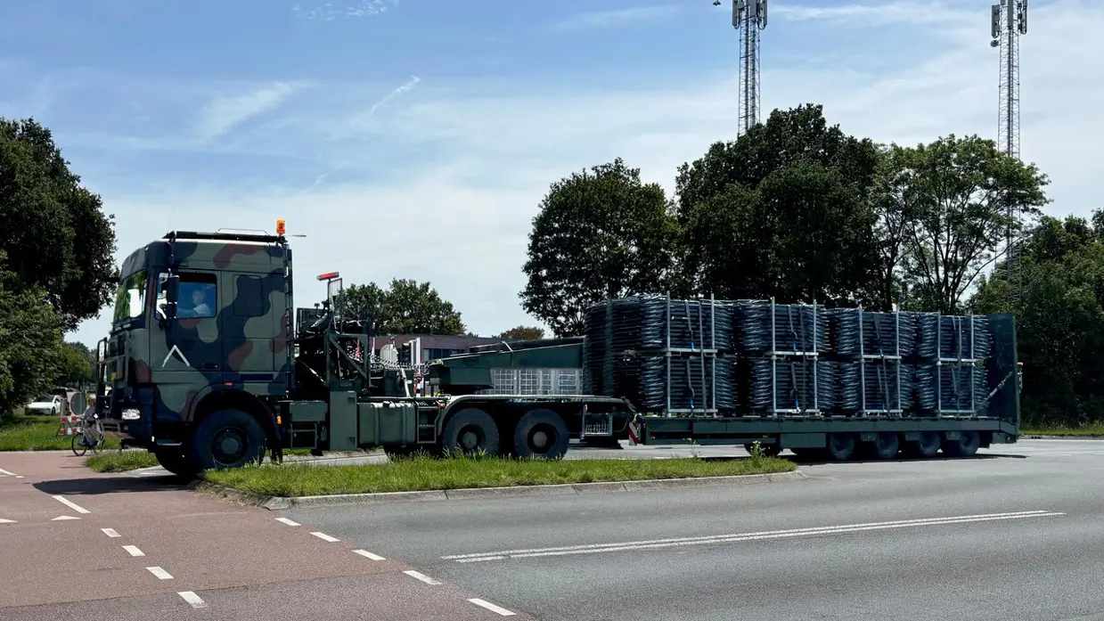 een groene trekker-oplegger beladen met veldbedden op een kruising.  