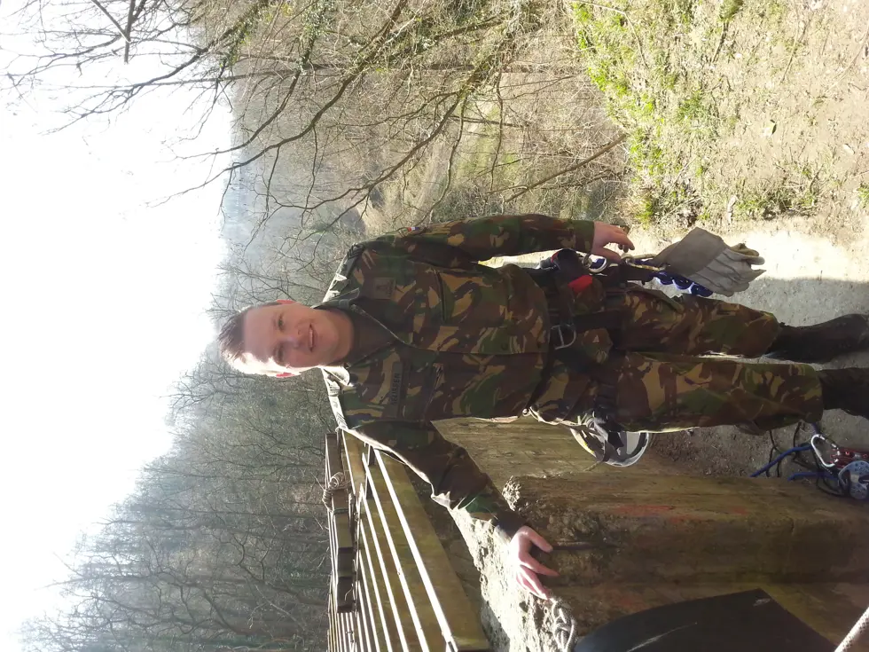 Een jonge Tim Nelissen in gevechtstenue poseert voor de foto in het buitenterrein. Om zijn heupen draagt hij een veiligheidstuigje met daaraan een helm en handschoenen vastgeklikt. Op de grond ligt een touw met karabijnhaak.
