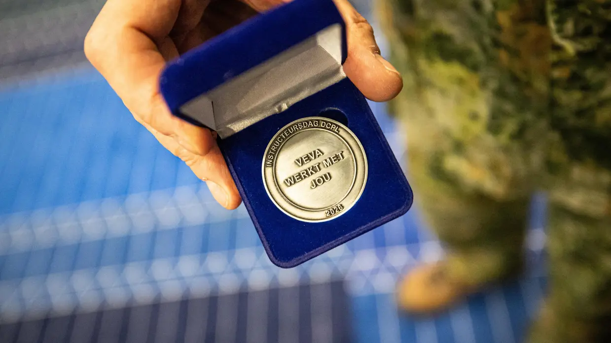 Een zilverkleurige munt met de tekst ‘Instructeursdag DCPL 2026; VeVa werkt met jou’. Deze wordt door een hand van een militair getoond in een blauw doosje.