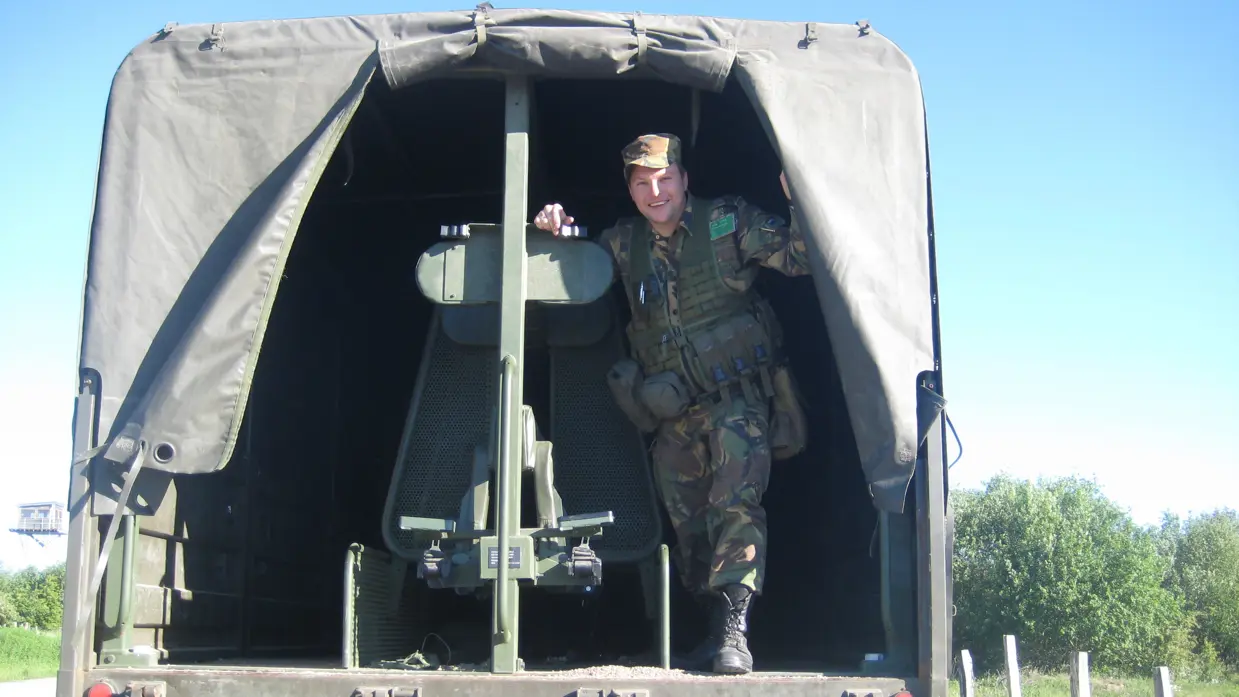 Een jonge Tim Nelissen in gevechtstenue poseert staand in de open achterkant van een militaire vrachtwagen en kijkt in de camera.