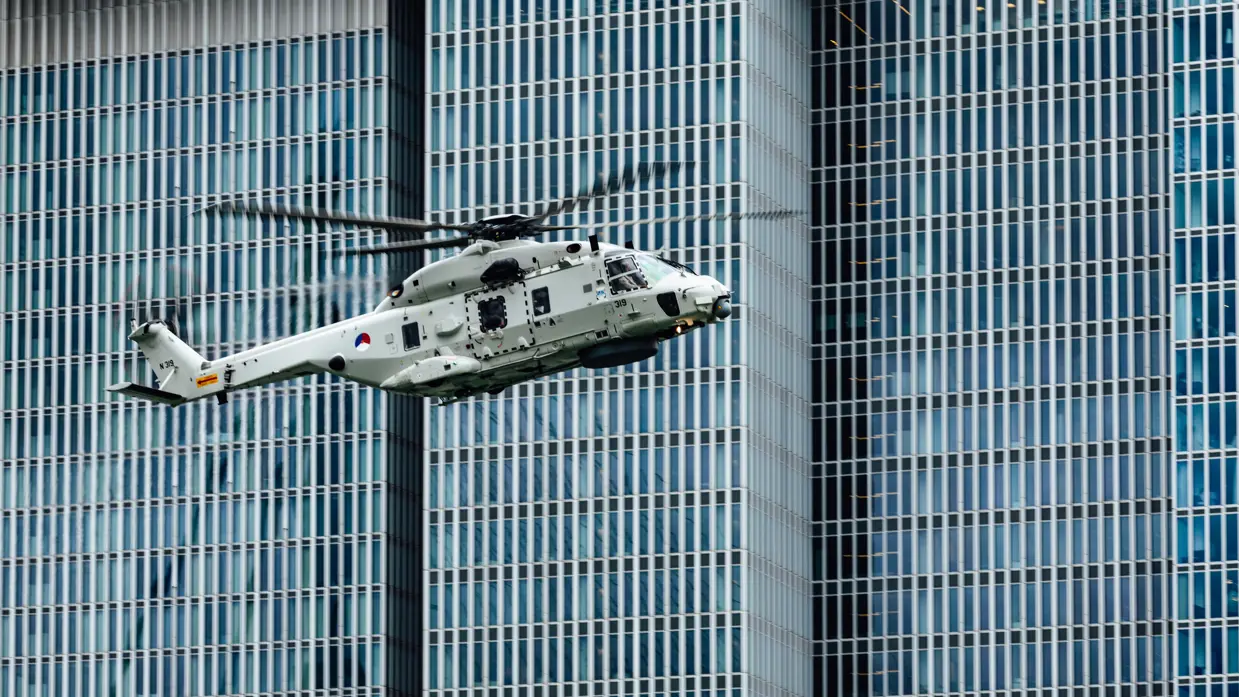 Een vliegende NH90 tegen een achtergrond van flatgebouwen.   