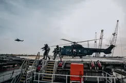Apache-gevechtshelikopter op platform. Militairen met getrokken wapens ervoor. Verder weg een Apache in de lucht.
