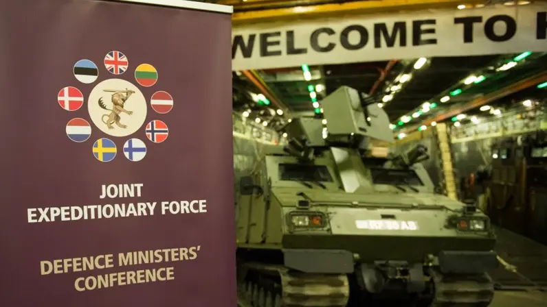 Een banner met daarop de tekst ‘Joint Expeditionary Force’ en de vlaggen van de deelnemende landen. Daarachter onscherp een militair voertuig.