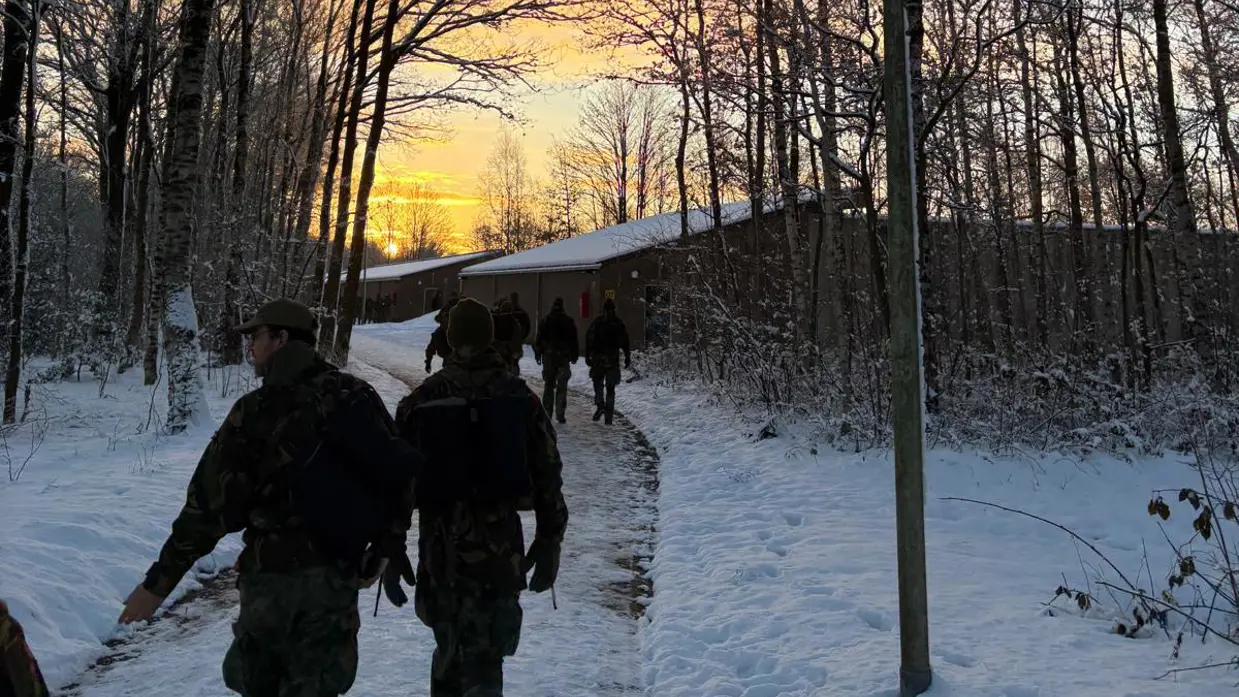 Deelnemers van de Nationale Weerbaarheidstraining lopen door een besneeuwd oefengebied tijdens zonsopkomst.