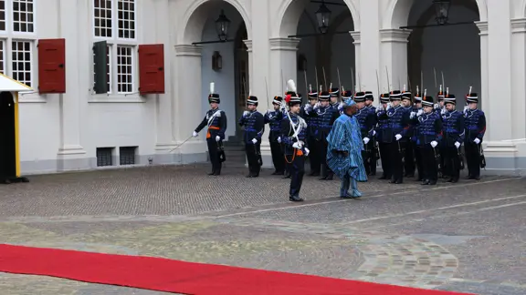 links: Een vrouw in een prachtige, versierde mantel loopt over een rode loper, terwijl een groep geüniformeerde wachten in blauwe tenues met sabel en rode pluim haar salueren. Midden: Een man in een blauwe, traditioneel gewaad loopt langs een rij wachten in blauwe uniformen met sabels en pluimen, voor een wit gebouw met rode luiken en een rode loper. Rechts: Een man in een donkere jas loopt langs een rij wachten in blauwe uniformen met pluimen en sabels, voor een wit gebouw met rode luiken en ramen.
