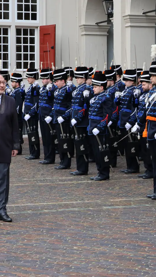 links: Een vrouw in een prachtige, versierde mantel loopt over een rode loper, terwijl een groep geüniformeerde wachten in blauwe tenues met sabel en rode pluim haar salueren. Midden: Een man in een blauwe, traditioneel gewaad loopt langs een rij wachten in blauwe uniformen met sabels en pluimen, voor een wit gebouw met rode luiken en een rode loper. Rechts: Een man in een donkere jas loopt langs een rij wachten in blauwe uniformen met pluimen en sabels, voor een wit gebouw met rode luiken en ramen.