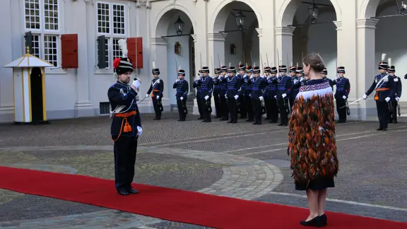 links: Een vrouw in een prachtige, versierde mantel loopt over een rode loper, terwijl een groep geüniformeerde wachten in blauwe tenues met sabel en rode pluim haar salueren. Midden: Een man in een blauwe, traditioneel gewaad loopt langs een rij wachten in blauwe uniformen met sabels en pluimen, voor een wit gebouw met rode luiken en een rode loper. Rechts: Een man in een donkere jas loopt langs een rij wachten in blauwe uniformen met pluimen en sabels, voor een wit gebouw met rode luiken en ramen.