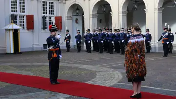 links: Een vrouw in een prachtige, versierde mantel loopt over een rode loper, terwijl een groep geüniformeerde wachten in blauwe tenues met sabel en rode pluim haar salueren. Midden: Een man in een blauwe, traditioneel gewaad loopt langs een rij wachten in blauwe uniformen met sabels en pluimen, voor een wit gebouw met rode luiken en een rode loper. Rechts: Een man in een donkere jas loopt langs een rij wachten in blauwe uniformen met pluimen en sabels, voor een wit gebouw met rode luiken en ramen.