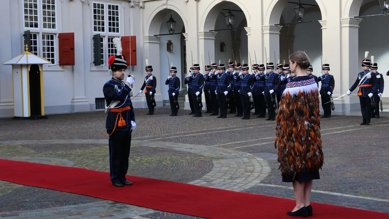 links: Een vrouw in een prachtige, versierde mantel loopt over een rode loper, terwijl een groep geüniformeerde wachten in blauwe tenues met sabel en rode pluim haar salueren. Midden: Een man in een blauwe, traditioneel gewaad loopt langs een rij wachten in blauwe uniformen met sabels en pluimen, voor een wit gebouw met rode luiken en een rode loper. Rechts: Een man in een donkere jas loopt langs een rij wachten in blauwe uniformen met pluimen en sabels, voor een wit gebouw met rode luiken en ramen.