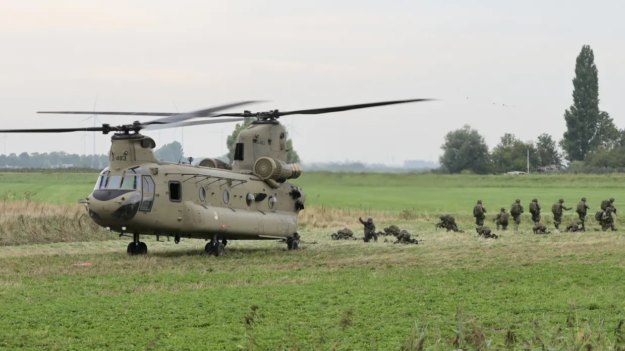 Militaire helikopter op een grasveld met soldaten in camouflage die uitstappen, omringd door groen landschap en bomen.